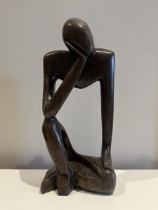 A wooden statue, shaped like man sitting with one leg crossed over the other, face resting on his hand, deep in thought.