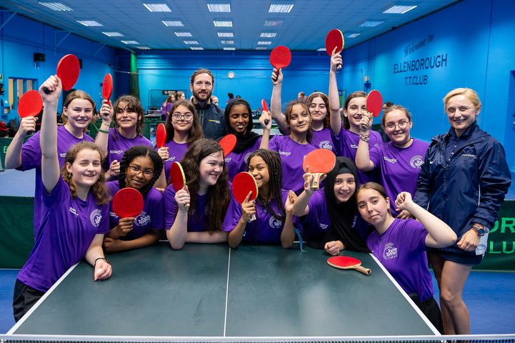 A group of young girls gathered together, smiling and holding table tennis paddles in front of a table tennis table