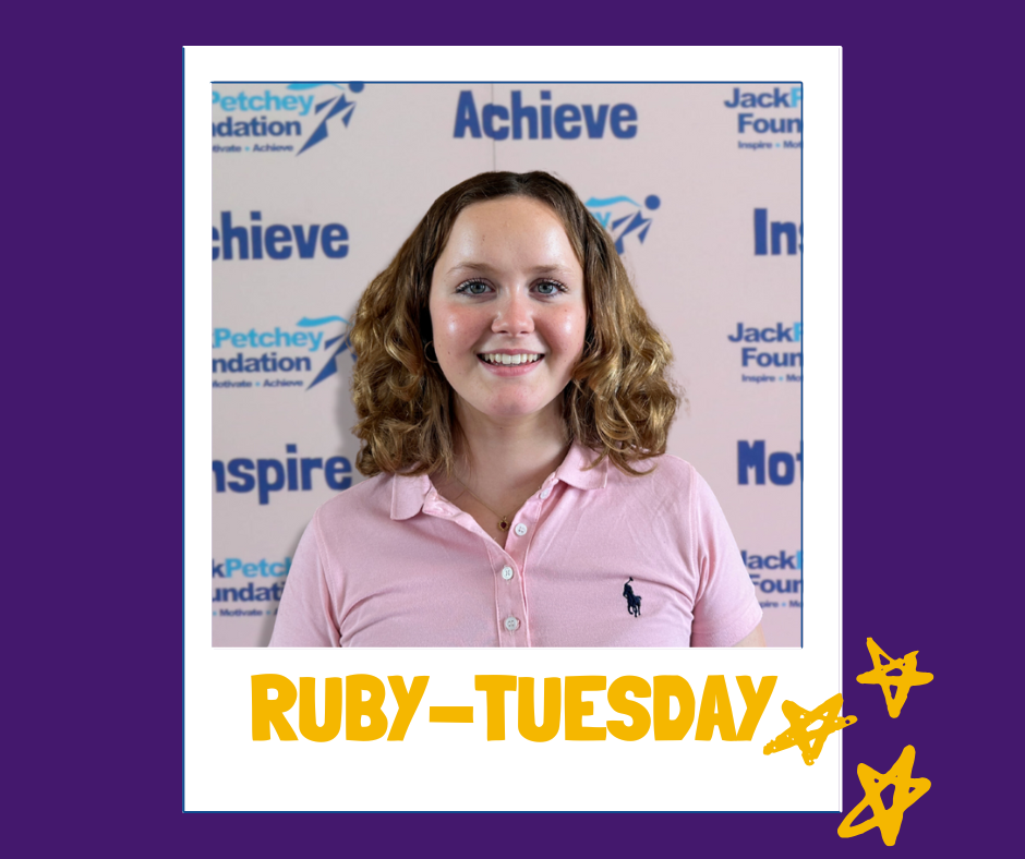 A portrait-style image of a person with shoulder-length curly blonde hair wearing a light pink collared shirt. They are standing in front of a branded backdrop featuring the Jack Petchey Foundation logo and words such as “Achieve,” “Inspire,” and “Motivate.” The photo is framed like a polaroid with a purple border, yellow star doodles in the bottom right corner, and the name “RUBY–TUESDAY” written in bold yellow letters at the bottom.