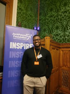 person stands indoors beside a tall blue banner for the Jack Petchey Foundation, printed with the word “INSPIRE” repeatedly. The person is wearing a black long‑sleeved top, light-coloured trousers, and an orange visitor lanyard. The background features ornate wooden panelling and decorative green patterned wallpaper.