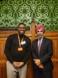 Two people stand side by side in a richly decorated room with ornate wooden panelling and patterned green wallpaper. One person wears a black top with a visitor lanyard, and the other is dressed in a suit with a tie and a turban. Both are posed facing the camera.