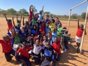 A large group of excited children posing playfully on a sandy field with their arms raised.
