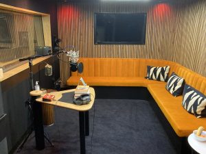 A modern recording studio with a curved orange upholstered bench along the back wall, decorated with patterned cushions. In front of the bench is a microphone setup on a stand, a small table with headphones and books, and acoustic wood paneling covering the walls. A large screen is mounted on the wall above the seating area, and studio equipment is arranged around the room.