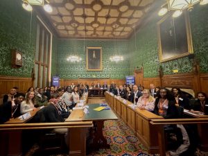 A grand room, with ornate green wall paper, big portraits and wooden panelling. There are lots of people in rows with Jack Petchey Foundation banners in the background that say 'inspire' and 'standing up for youth voice'.