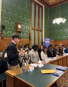 A grand room, with ornate green wall paper, big portraits and wooden panelling. There is a Jack Petchey Foundation banner in the background that repeats the word 'inspire'. A young person is stood up speaking whilst those sat down look at him.