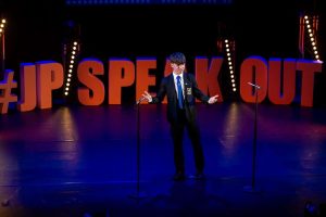 A big stage with red letters that say '#JP SPEAK OUT' around the back of the stage. There is blue lighting and a young person in between two microphones speaking.