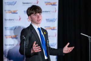 A young person is speaking between two microphones, looking out strongly. There is a banner behind him with Jack Petchey Foundation, Jack Petchey's Speak Out Challenge and Speakers Trust logos.