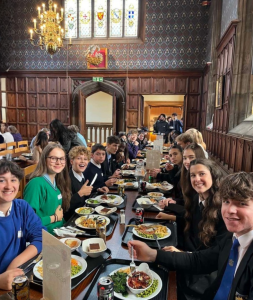 A grand room, with ornate wall paper and wooden panelling. There are young people sat at a long table earing food.
