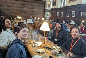 A group of young people seated around a long wooden table inside a historic hall, with portraits on the walls, eating and talking together under table lamps.