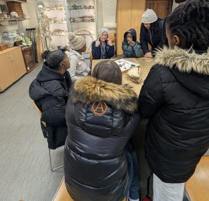 A small group of young people gathered around a table, examining objects while an adult facilitator gestures and leads a discussion.
