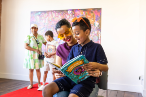 A young person wearing orange star-shaped glasses sits on a chair reading a colourful book titled Alien World and Other Stories with an adult seated behind them. Both are looking at the open book. A red carpet covers part of the wooden floor in the bright indoor space.