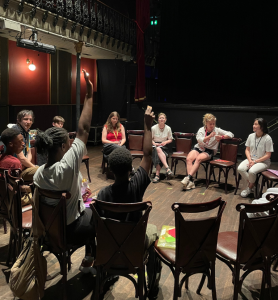 A group workshop takes place in a small theatre auditorium, with participants seated in a circle of chairs on a wooden floor. Two participants in the foreground have their hands raised while several facilitators and attendees sit around the circle. The venue features ornate balcony railings, red walls, stage curtains, and dim theatrical lighting, creating an intimate discussion setting.