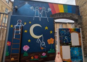A colourful mural painted on an outdoor wall beneath a brick archway, featuring illustrated figures, stars, flowers, a crescent moon and a rainbow. One figure climbs a ladder towards the sky, while another hangs from the moon holding a watering can. In the foreground, a hand holds up an original artwork or design template in front of the completed mural, showing how the creative concept was transformed into a large-scale piece of public art. The scene is set in an urban courtyard between brick buildings.