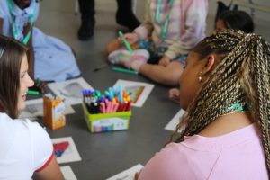 A group of young people sit in a circle on the floor during a creative workshop, using coloured pens and paper for drawing activities. A container of brightly coloured markers is placed in the centre, with artwork and stationery spread around the group. Two participants are visible in the foreground, while others sit around them in an indoor classroom or community setting.